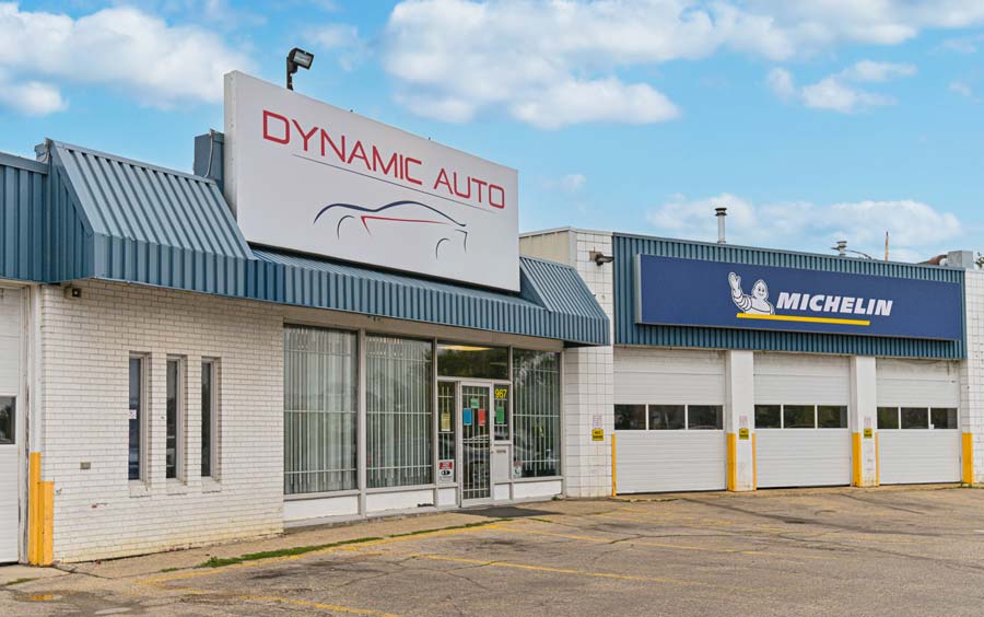 Front view of Dynamic Auto, featuring a prominent sign above the entrance, located next to a Michelin service area. The building has a clean, industrial design with three large garage doors and clear blue skies above.